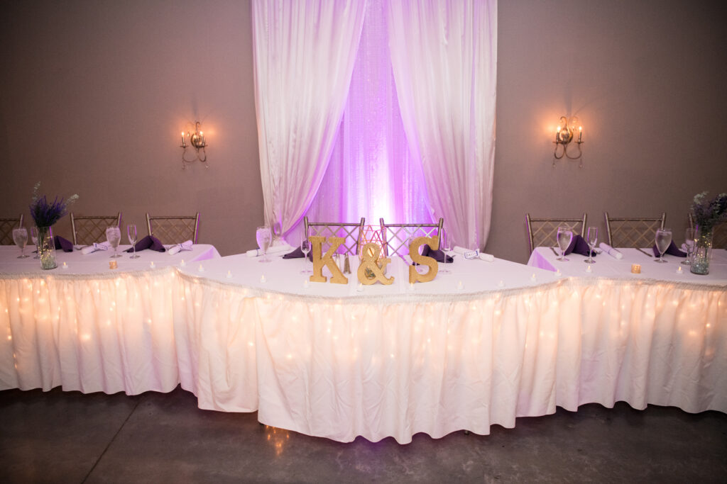 A beautifully set wedding reception table adorned with a white tablecloth and illuminated by soft purple lights.